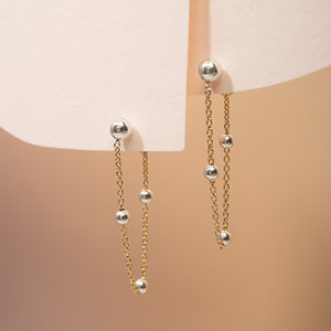 A pair of gold and silver dangle earrings with small spherical beads, displayed on a minimalist cream-colored jewelry stand against a warm beige background.
