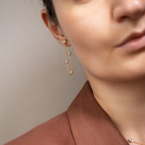 A close-up of a model wearing a delicate gold dangle earring with small pearl-like beads, paired with a brown blazer.