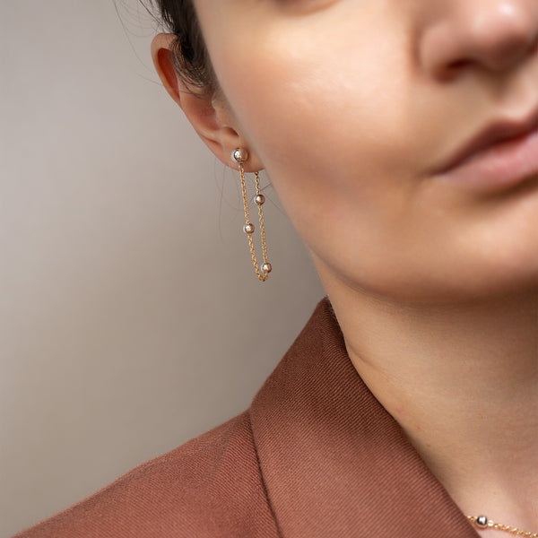 A close-up of a model wearing a delicate gold dangle earring with small pearl-like beads, paired with a brown blazer.