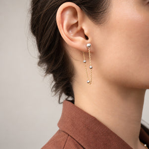 A close-up of a model's ear wearing a delicate gold dangle earring with small silver-toned beads and pearl-like accents, paired with a brown collared top against a soft neutral background.