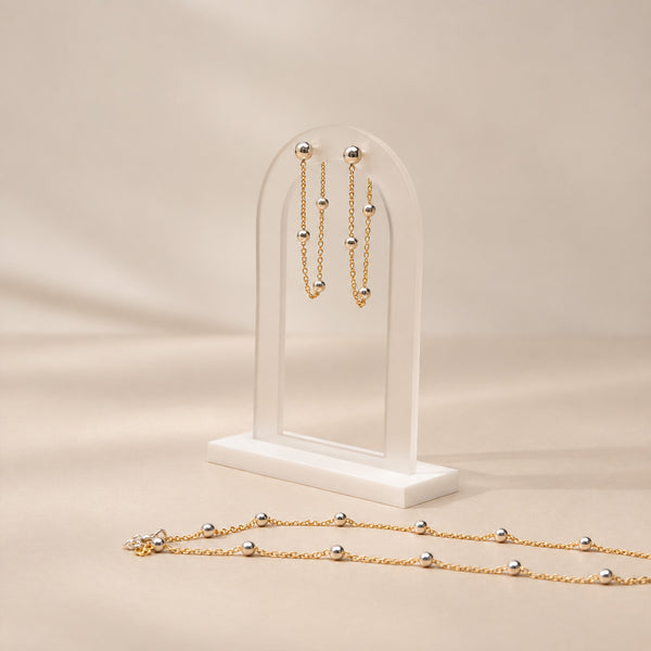Gold beaded jewelry set displayed on a clear acrylic stand, featuring matching drop earrings and a long necklace, photographed on a warm beige background.