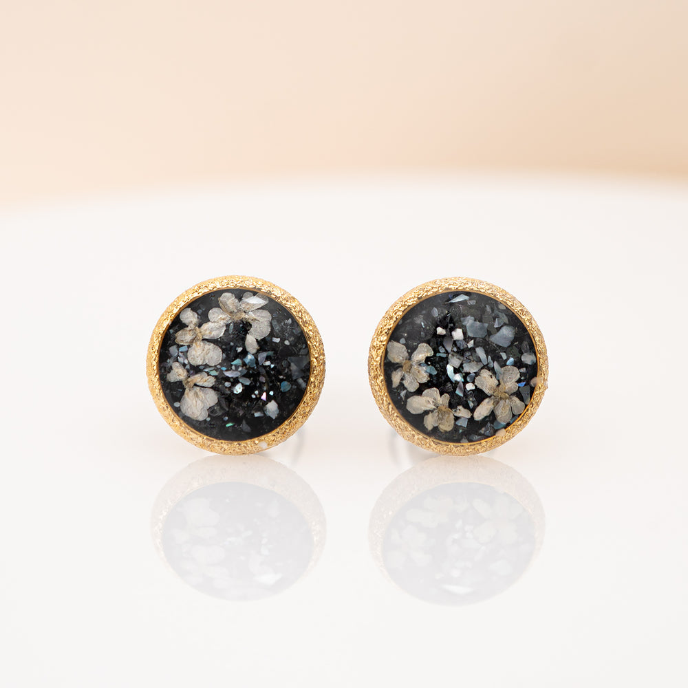 A pair of round gold-tone stud earrings featuring a black druzy stone inlay with white and gray crystalline patterns, set against a clean off-white background.
