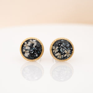 A pair of round stud earrings with textured gold borders and dark gray to black druzy stone centers containing white and gray crystalline flecks, displayed on a reflective white surface against a soft beige background.