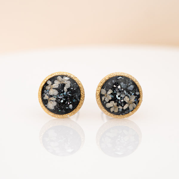 A pair of round stud earrings with textured gold borders and dark gray to black druzy stone centers containing white and gray crystalline flecks, displayed on a reflective white surface against a soft beige background.