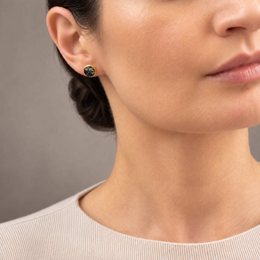 A pair of round gold-tone stud earrings featuring a black druzy stone inlay with white and gray crystalline patterns, set against a clean off-white background.