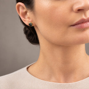 Close-up of a woman wearing a small round stud earring with a dark green stone and gold bezel setting, against a neutral gray background.