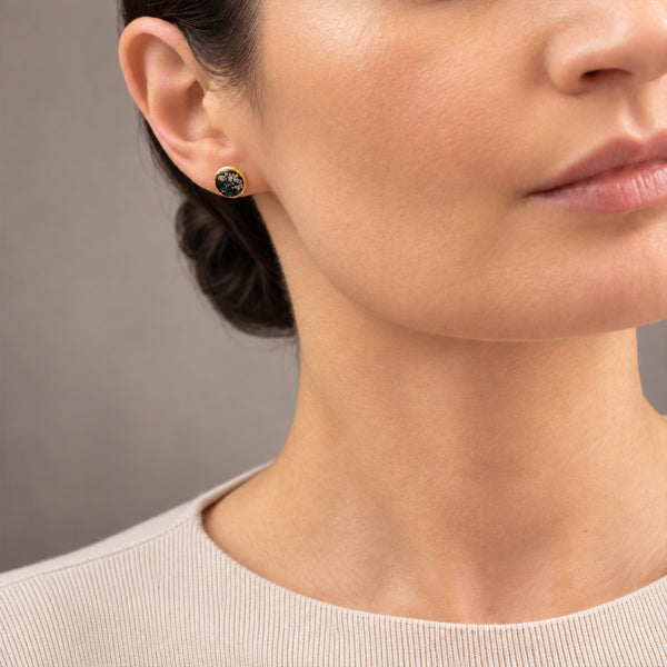 Close-up of a woman wearing a small round stud earring with a dark green stone and gold bezel setting, against a neutral gray background.