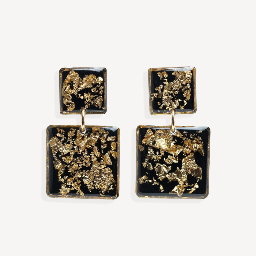 A pair of square drop earrings with textured gold leaf inlay on a black background, suspended from small gold rings against a light gray studio backdrop.