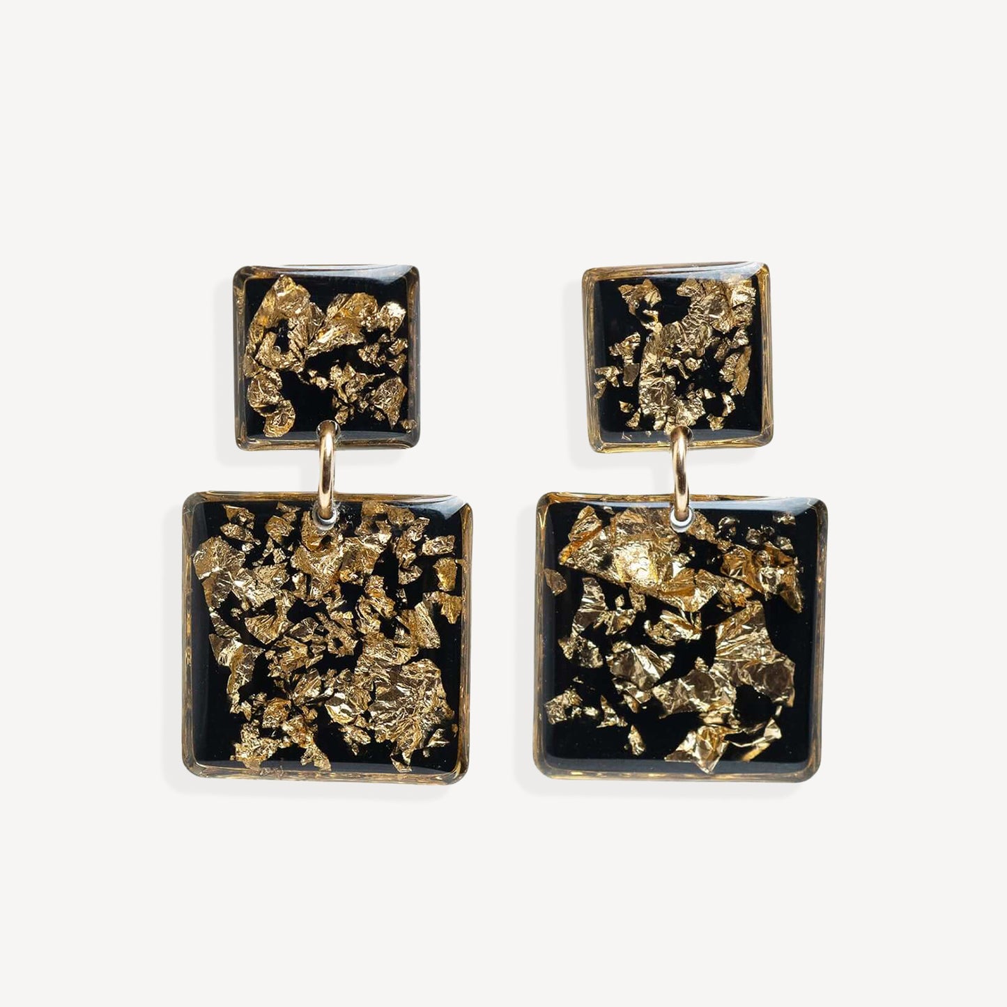 A pair of square drop earrings with textured gold leaf inlay on a black background, suspended from small gold rings against a light gray studio backdrop.
