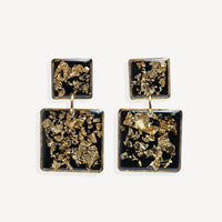 A pair of square drop earrings with textured gold leaf inlay on a black background, suspended from small gold rings against a light gray studio backdrop.