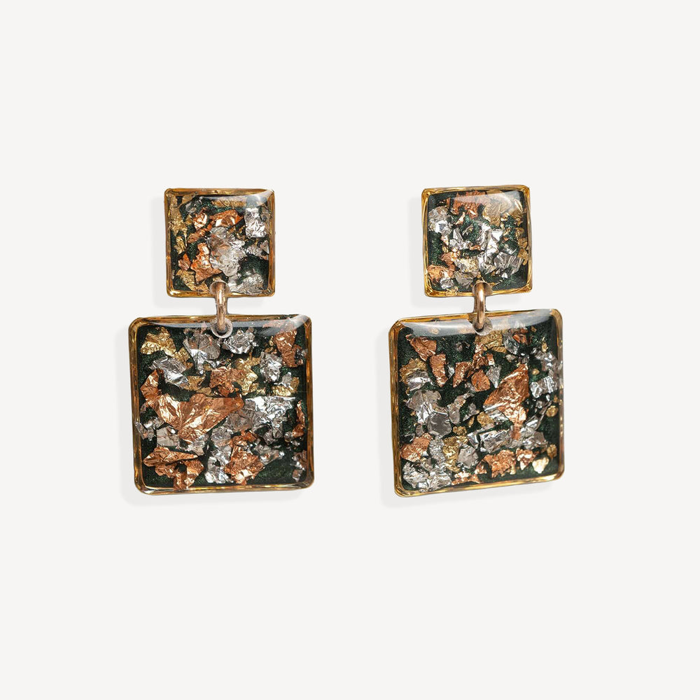 A pair of square drop earrings with textured, metallic inlay details in copper, silver, and dark green tones, set in a gold-tone frame on a light gray background.