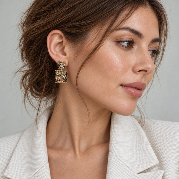 A close-up of a woman wearing a pair of square gold-toned earrings with colorful floral enamel detailing, paired with a light-colored blazer.