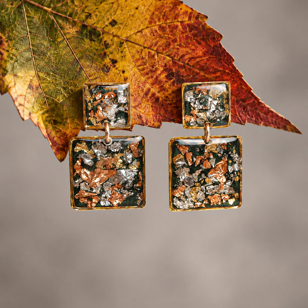 A pair of square drop earrings with textured, metallic inlay details in copper, silver, and dark green tones, set in a gold-tone frame on a light gray background.