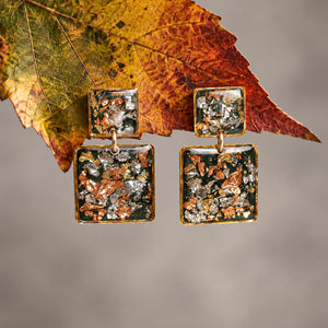 A pair of square drop earrings with textured metallic and earthy inlay details, hanging against a soft gray background with a large autumn leaf behind them.