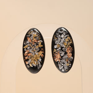 A pair of oval black earrings with embedded metallic flakes in silver, gold, and copper tones, displayed on a clear stand against a warm beige background.