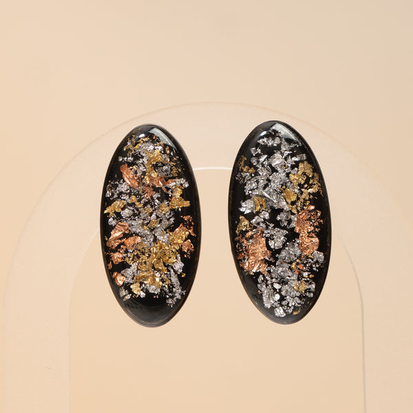 A pair of oval black earrings with embedded metallic flakes in silver, gold, and copper tones, displayed on a clear stand against a warm beige background.