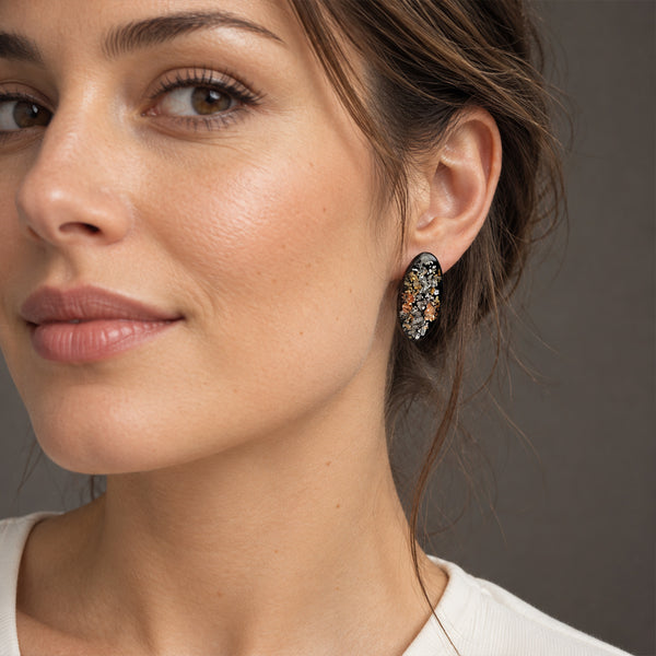 A close-up of a woman wearing a small, oval, multi-colored beaded earring in a studio setting with a neutral gray background.