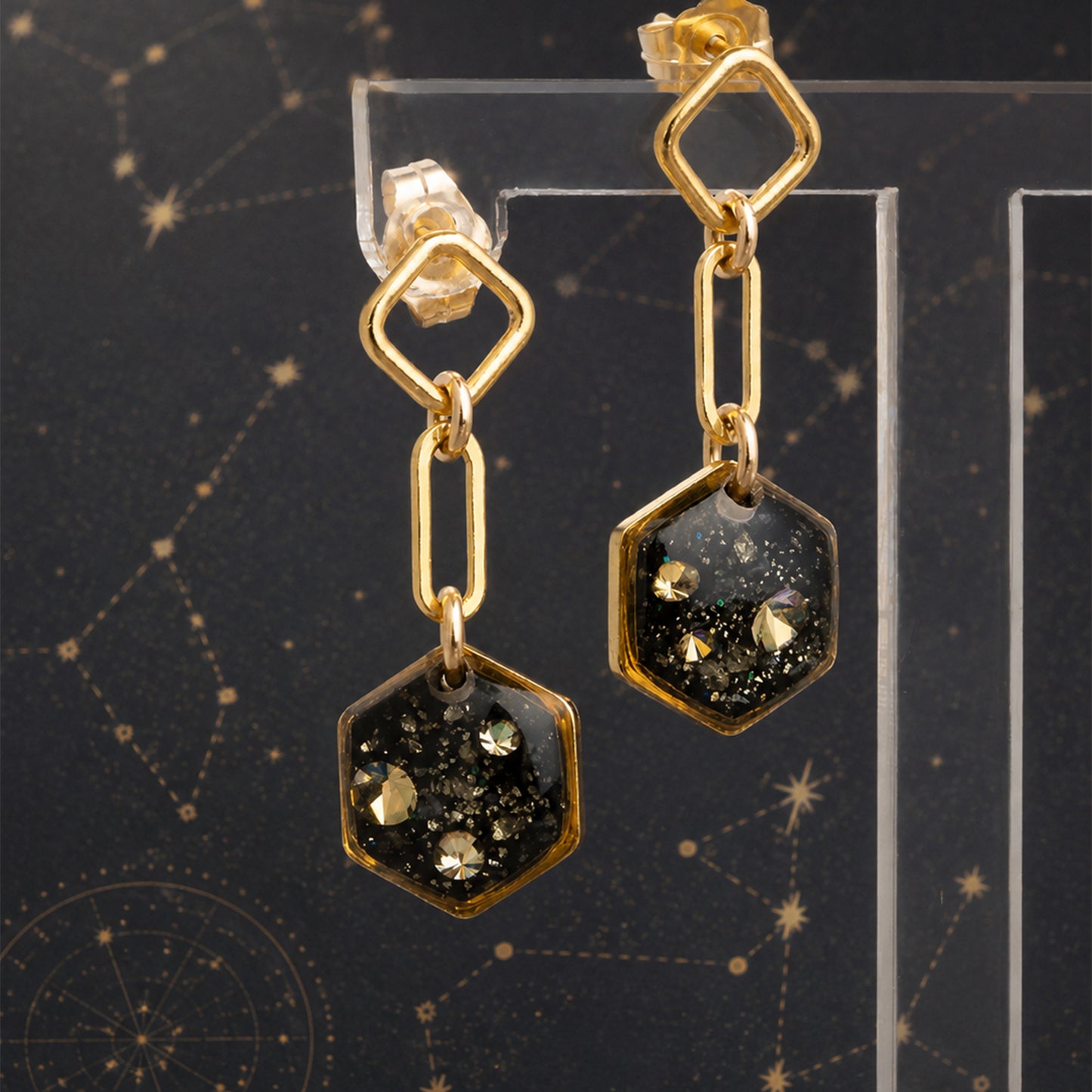 A pair of gold-toned dangle earrings with hexagonal charms filled with black glitter and small gold flecks, displayed on a clear acrylic stand against a dark starry background.