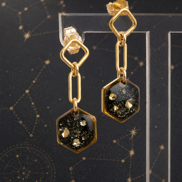 A pair of gold-toned dangle earrings with hexagonal charms filled with black glitter and small gold flecks, displayed on a clear acrylic stand against a dark starry background.