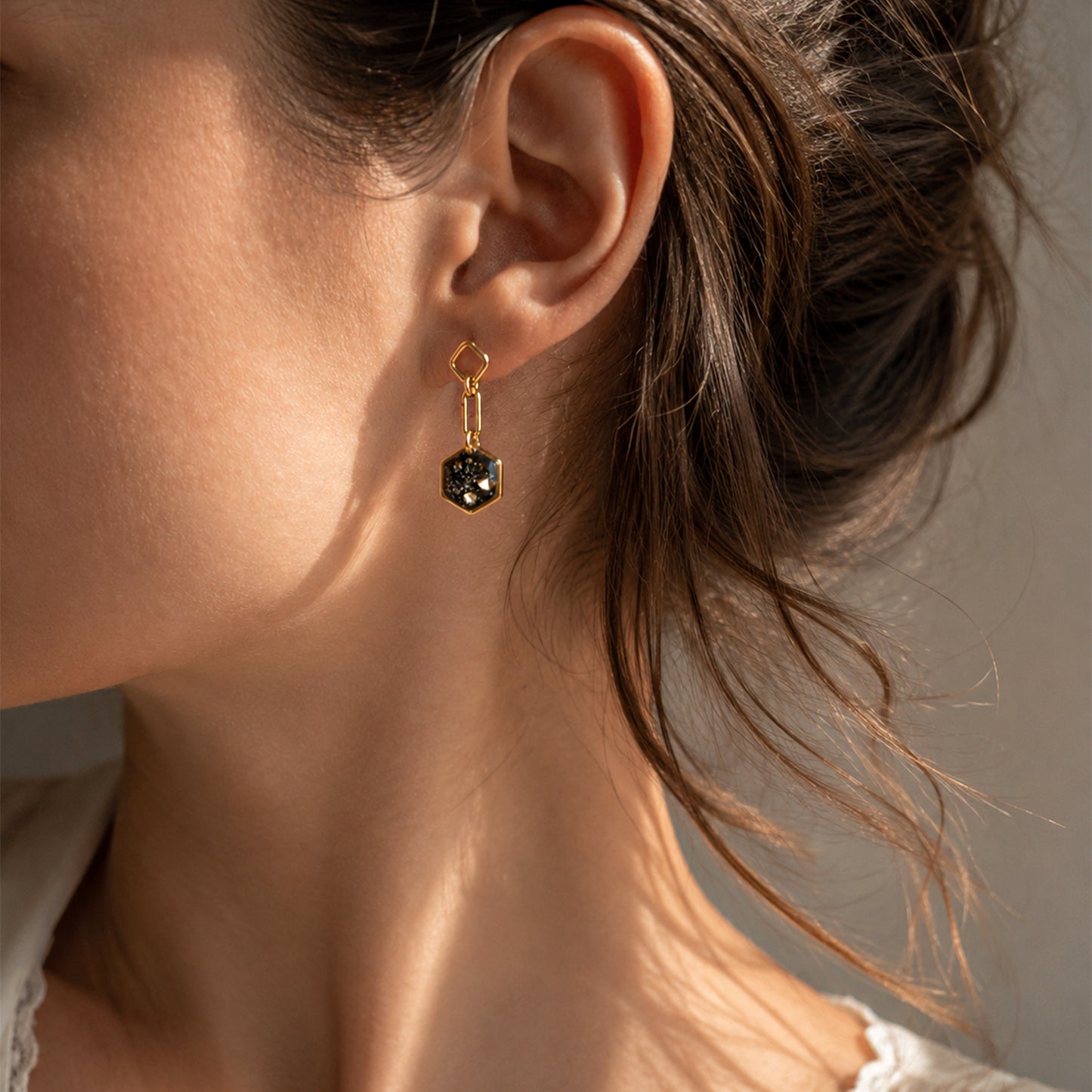 A close-up of a woman's ear wearing a delicate gold dangle earring with a faceted dark gemstone.