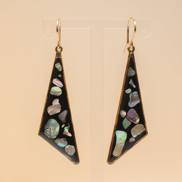 A pair of long triangular dangle earrings with iridescent abalone shell inlay on a black background and gold-tone hooks, displayed on clear acrylic stands against a warm beige backdrop.