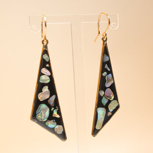 A pair of gold-tone dangle earrings with irregular triangular shapes, featuring a black background inlaid with iridescent abalone shell pieces, displayed on clear acrylic stands against a warm beige background.