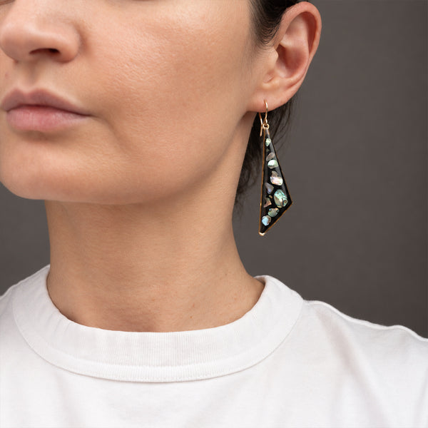 A close-up of a model wearing a long, triangular gold dangle earring decorated with iridescent abalone shell pieces. The earring features a gold-toned metal frame and hooks, with the shell inlays displaying shades of green, blue, and pink.