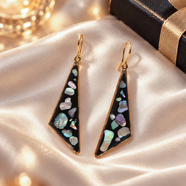 A pair of gold-tone drop earrings with triangular black inlays featuring iridescent mother-of-pearl shell pieces, displayed on a cream satin fabric next to a black gift box with a gold ribbon.