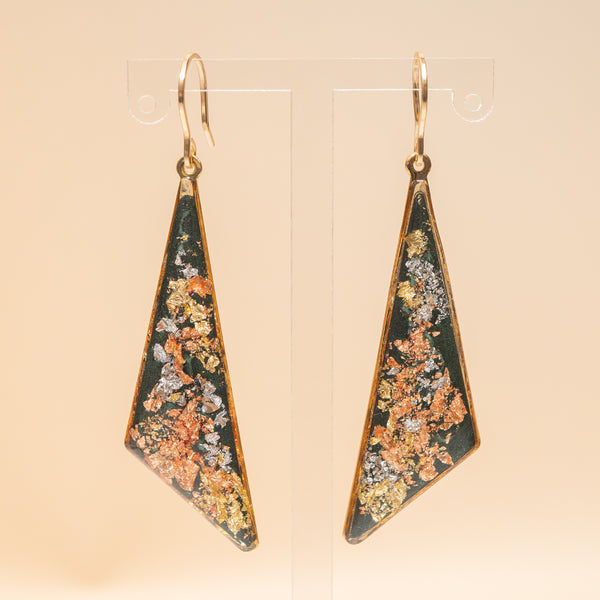 A pair of gold-tone dangle earrings with triangular resin pendants featuring a textured, metallic gold, copper, and silver floral pattern on a dark green background, displayed on clear acrylic stands.