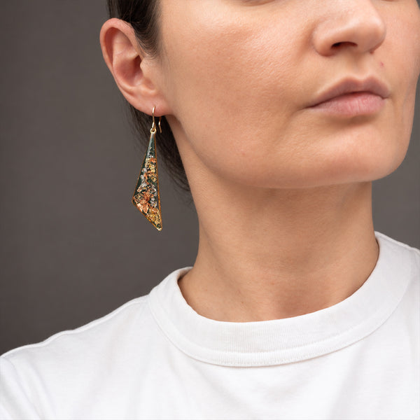 A close-up of a woman wearing a dangling triangular earring with a gold-tone frame and multicolored mosaic inlay, paired with a white crew-neck top against a dark gray background.