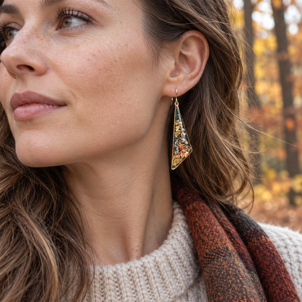A close-up lifestyle shot of a woman wearing a pair of triangular gold-tone earrings with colorful, textured inlay details, set against a blurred autumn forest background.