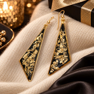 A pair of gold-tone geometric drop earrings with triangular shapes, featuring a black background filled with shimmering gold leaf flakes, displayed on a cream knit fabric with dark satin and gift packaging in the background.