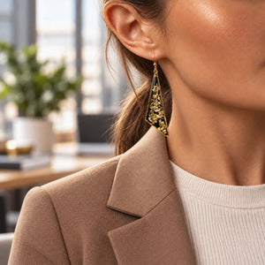 A close-up lifestyle shot of a woman wearing a gold triangular dangle earring with textured gold leaf detailing and a dark blue inlay, styled with a beige blazer and white ribbed top.
