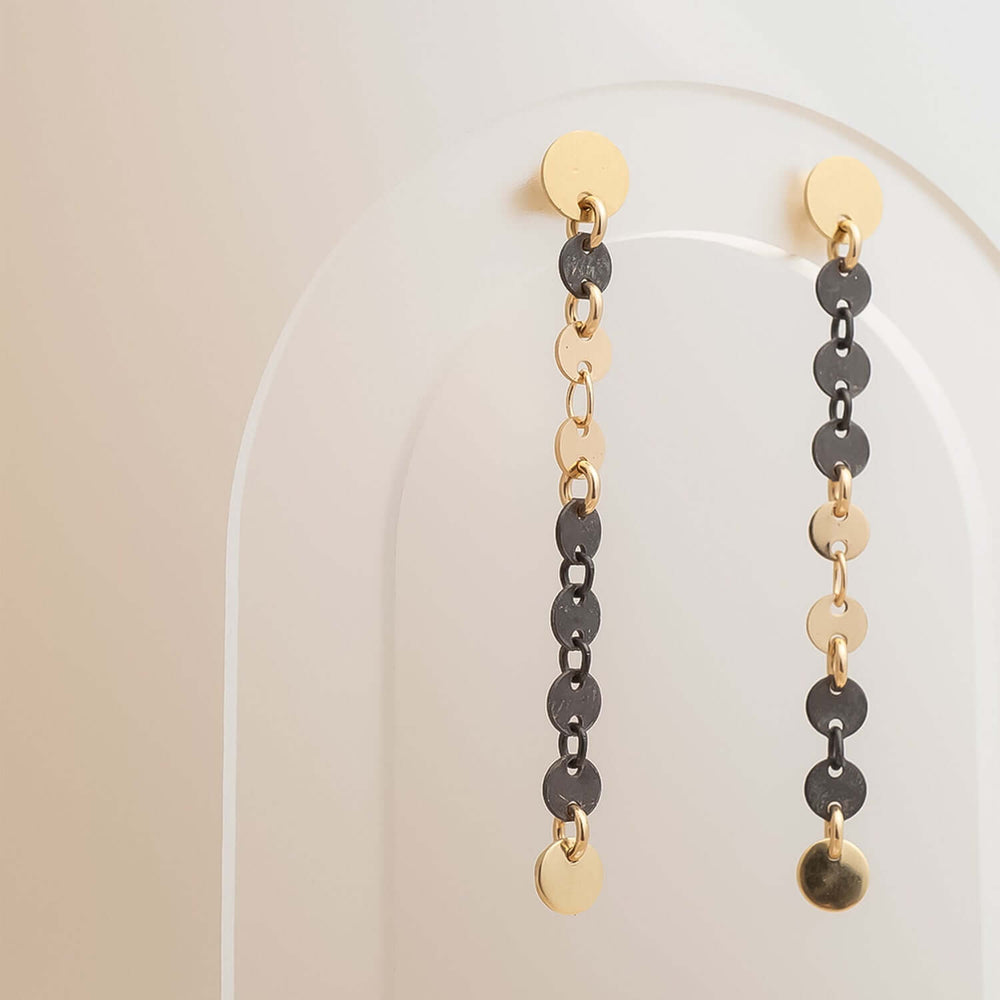 
                      
                        Pair of gold and black chain earrings on a clear stand against a beige background
                      
                    