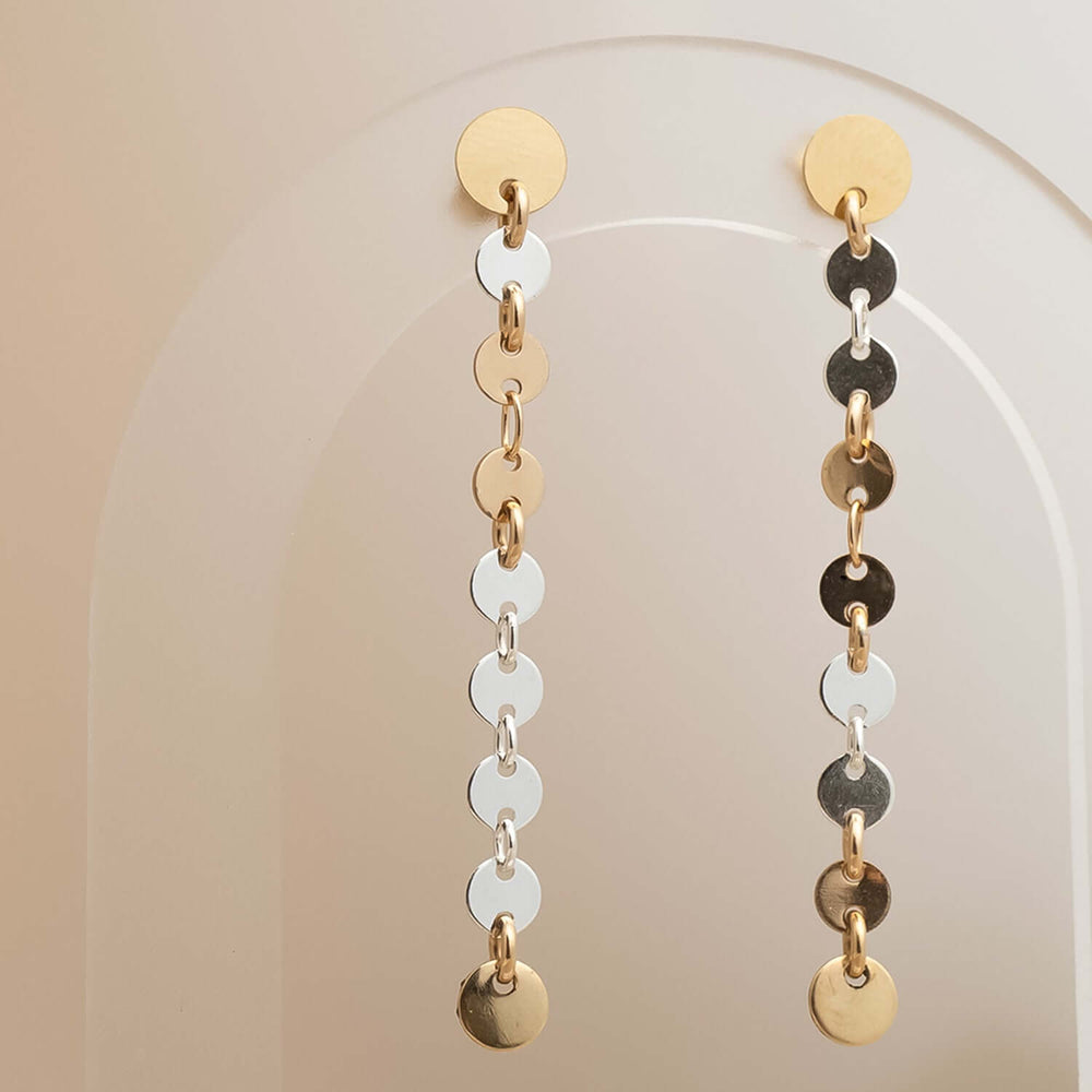 
                      
                        Pair of dangling earrings with gold, silver, and black circular pendants on a neutral background.
                      
                    