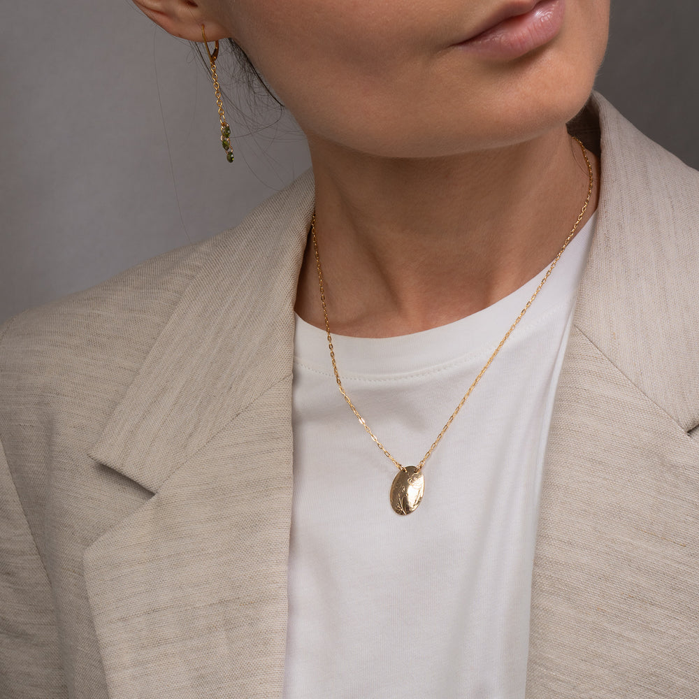 A gold oval pendant necklace with engraved floral details, suspended from a delicate gold chain against a light gray background.