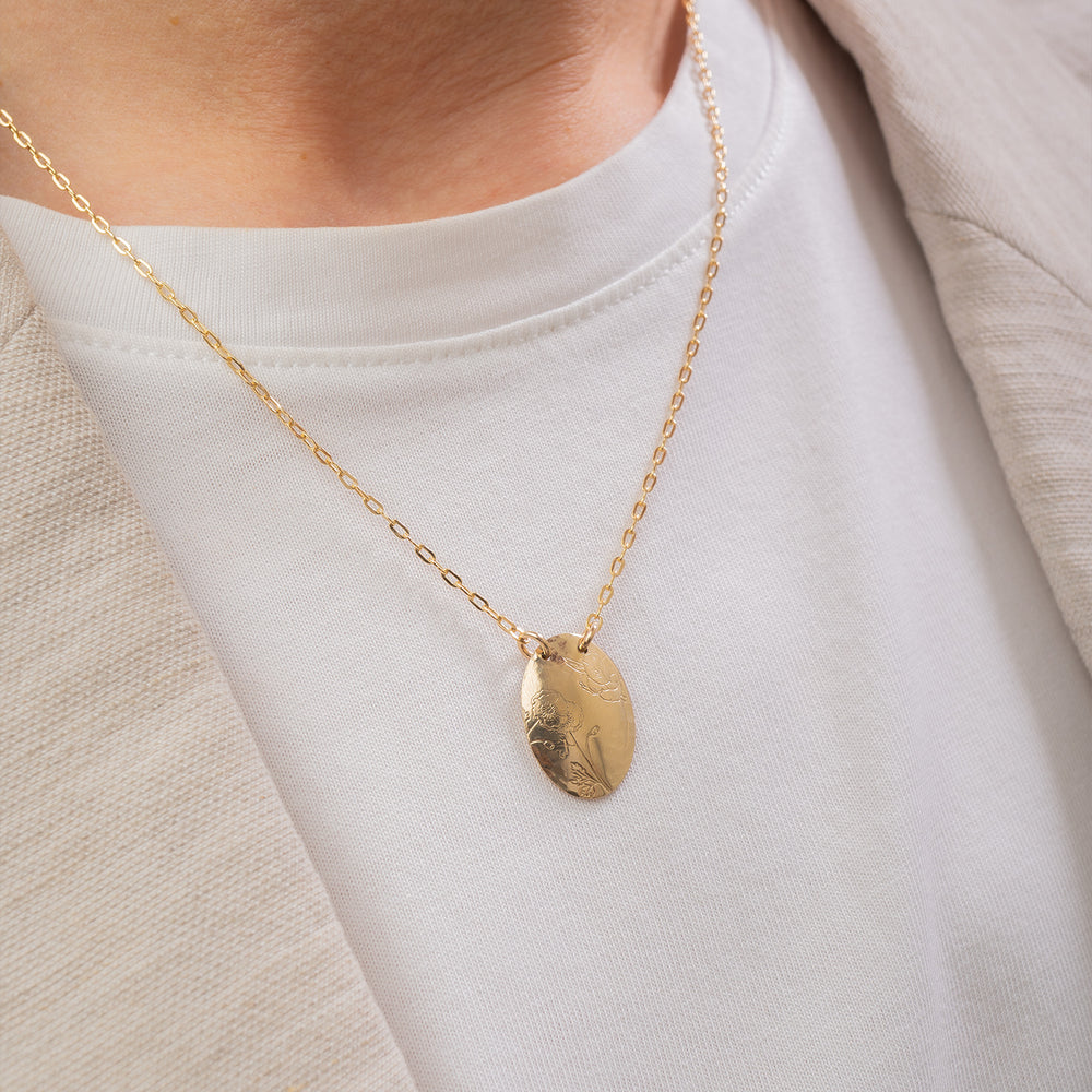 A gold oval pendant necklace with engraved floral details, suspended from a delicate gold chain against a light gray background.