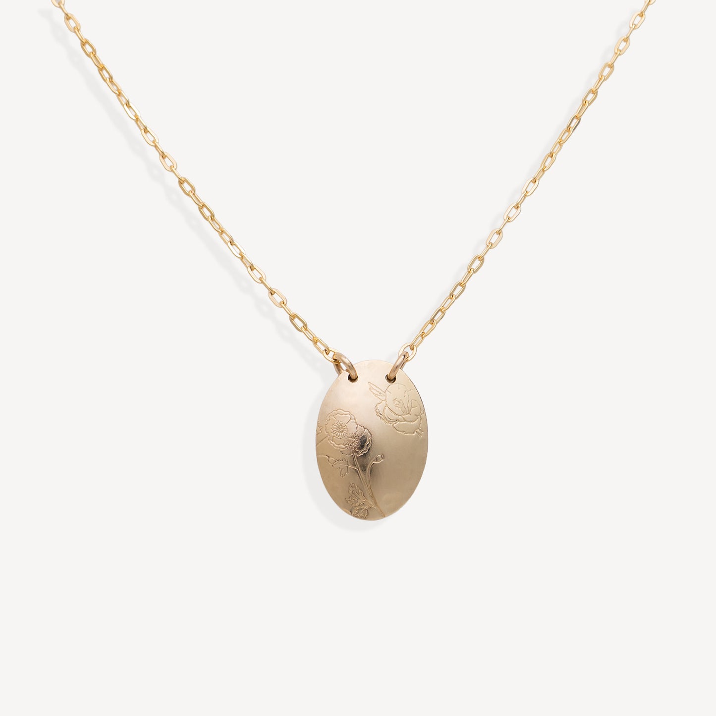 A delicate gold chain necklace with an oval pendant featuring an engraved floral design, displayed against a clean off-white background.