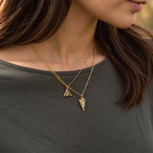 A close-up of a woman wearing a gold necklace with two triangular pendants, one featuring a textured stone and the other a smaller polished stone, over a dark gray t-shirt.