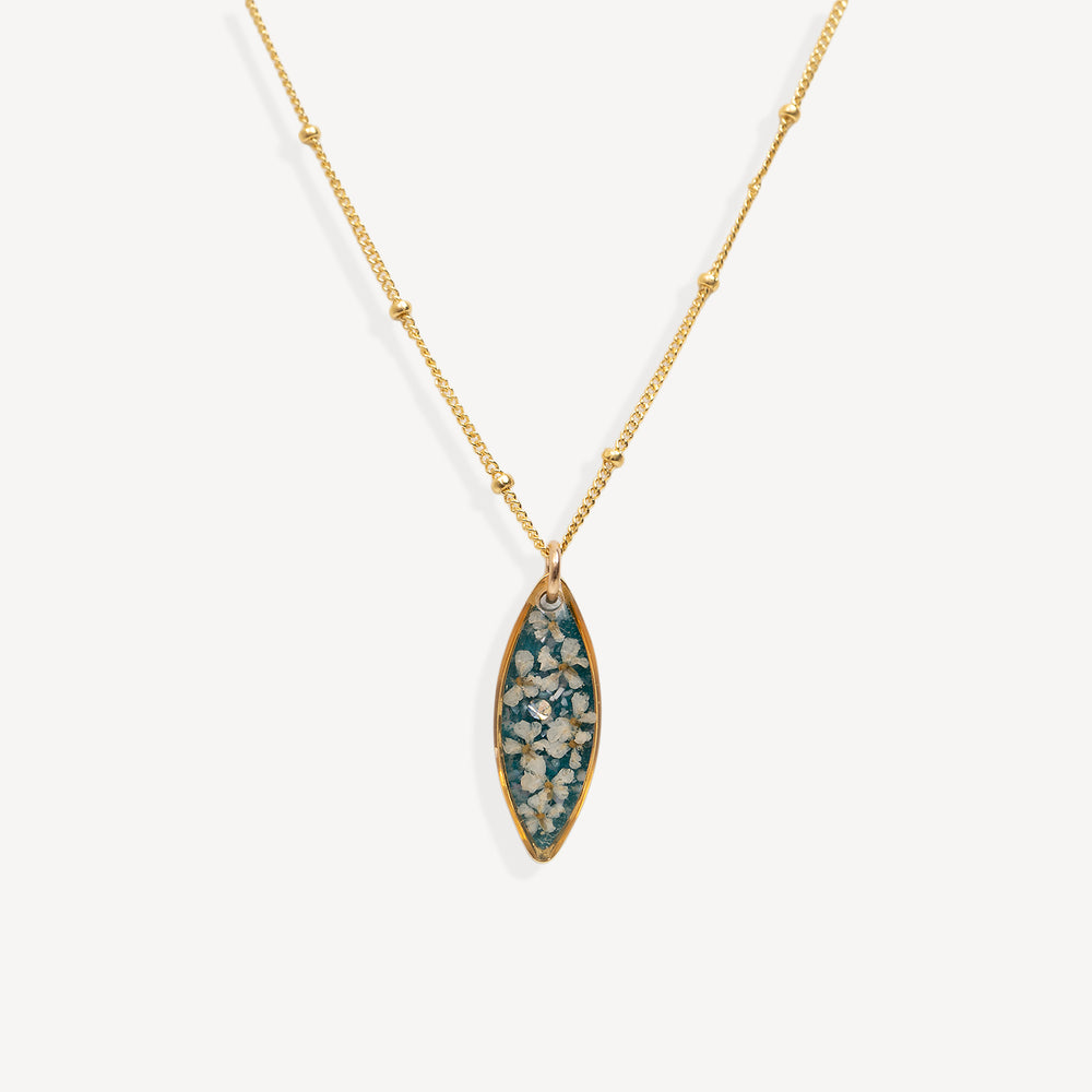 A gold chain necklace with a marquise-shaped pendant featuring a teal blue background and white floral details, shown against a light gray background.