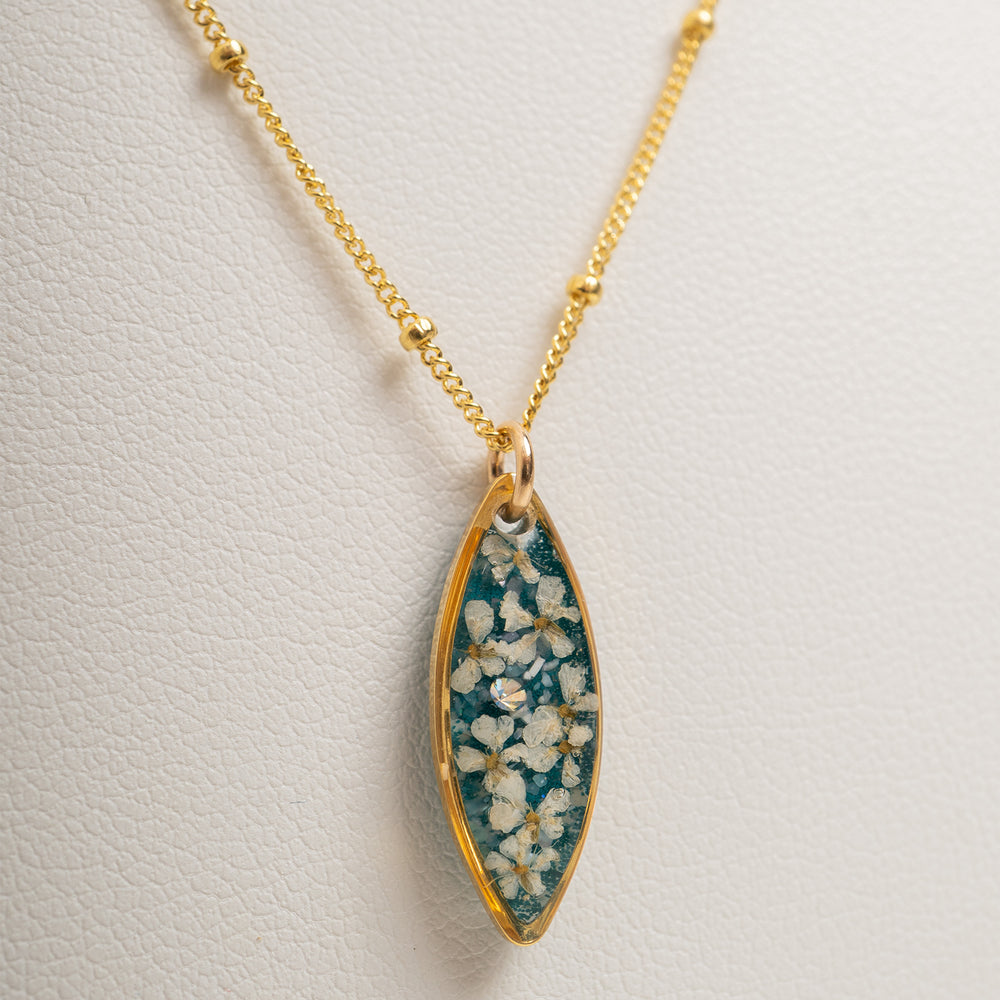A gold chain necklace with a marquise-shaped pendant featuring a teal blue background and white floral details, shown against a light gray background.
