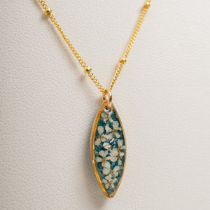 Gold chain necklace with an elongated marquise-shaped pendant featuring a teal blue background filled with small white flowers, displayed on a textured off-white surface.