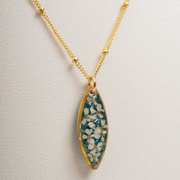 Gold chain necklace with an elongated marquise-shaped pendant featuring a teal blue background filled with small white flowers, displayed on a textured off-white surface.