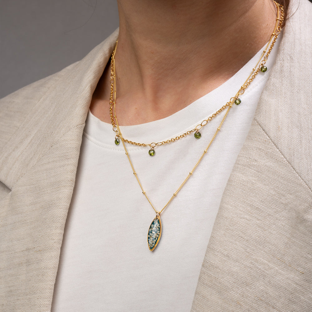 A gold chain necklace with a marquise-shaped pendant featuring a teal blue background and white floral details, shown against a light gray background.