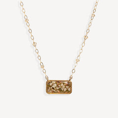 A gold chain necklace featuring a rectangular pendant filled with dried flowers and small botanical pieces, shown against a plain off-white background.