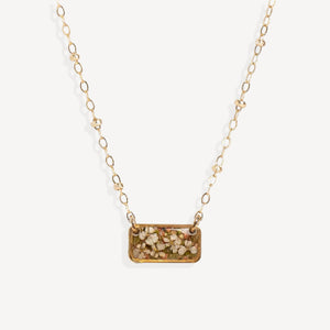 A gold chain necklace featuring a rectangular pendant filled with dried flowers and small botanical pieces, shown against a plain off-white background.