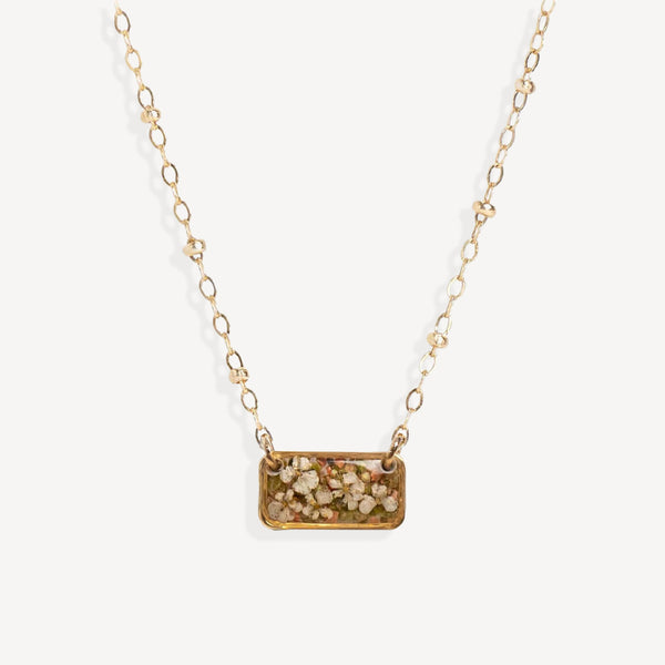 A gold chain necklace featuring a rectangular pendant filled with dried flowers and small botanical pieces, shown against a plain off-white background.