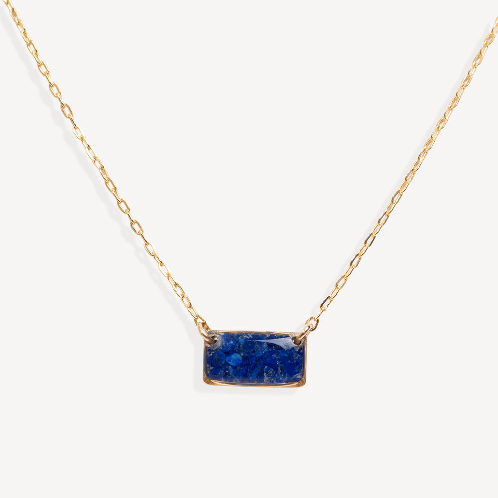 A delicate gold chain necklace featuring a small, rectangular blue stone pendant with gold bezel settings, shown against a clean off-white background.