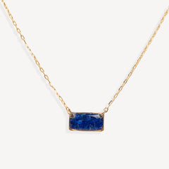 A delicate gold chain necklace featuring a small, rectangular blue stone pendant with gold bezel settings, shown against a clean off-white background.