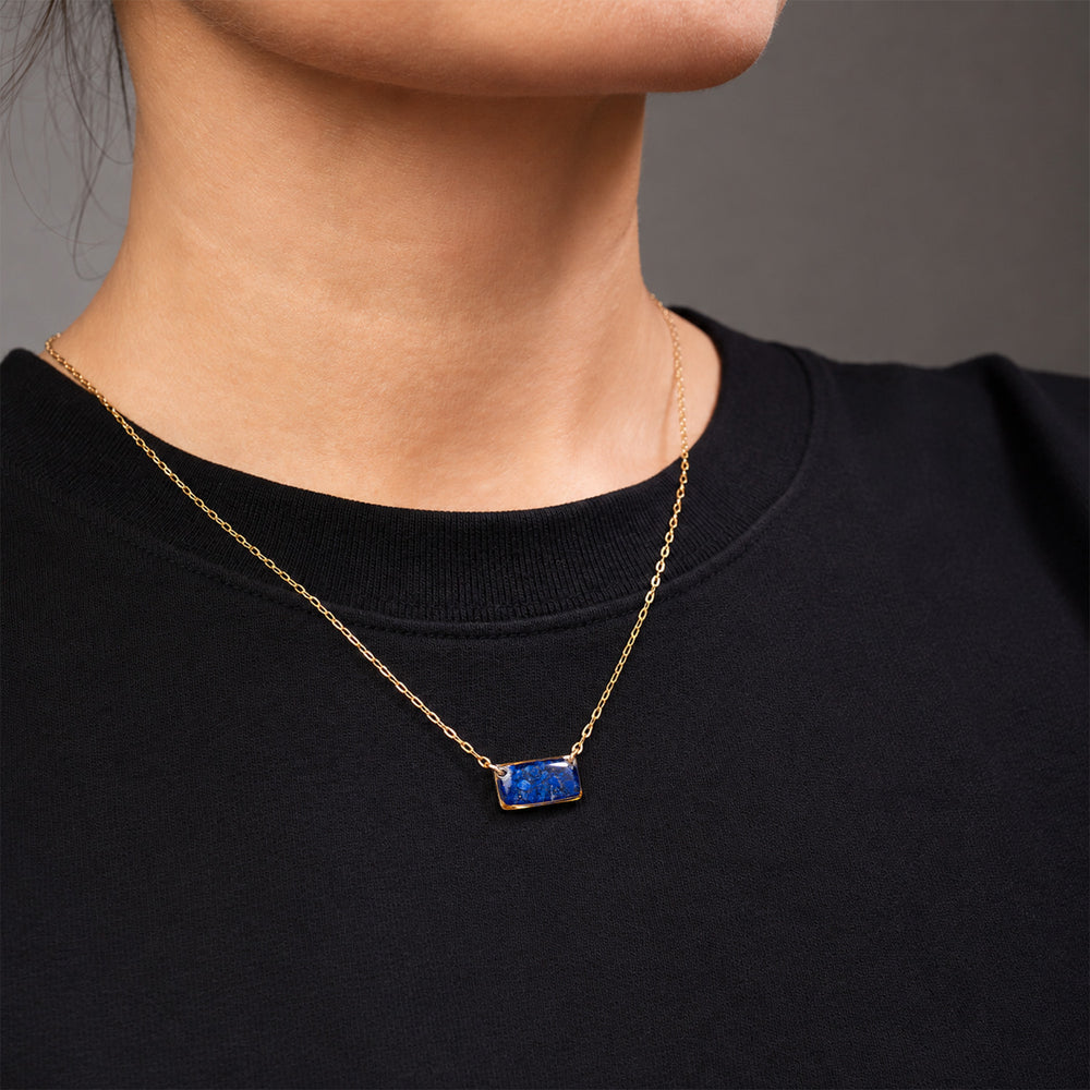 A delicate gold chain necklace featuring a small, rectangular blue stone pendant with gold bezel settings, shown against a clean off-white background.